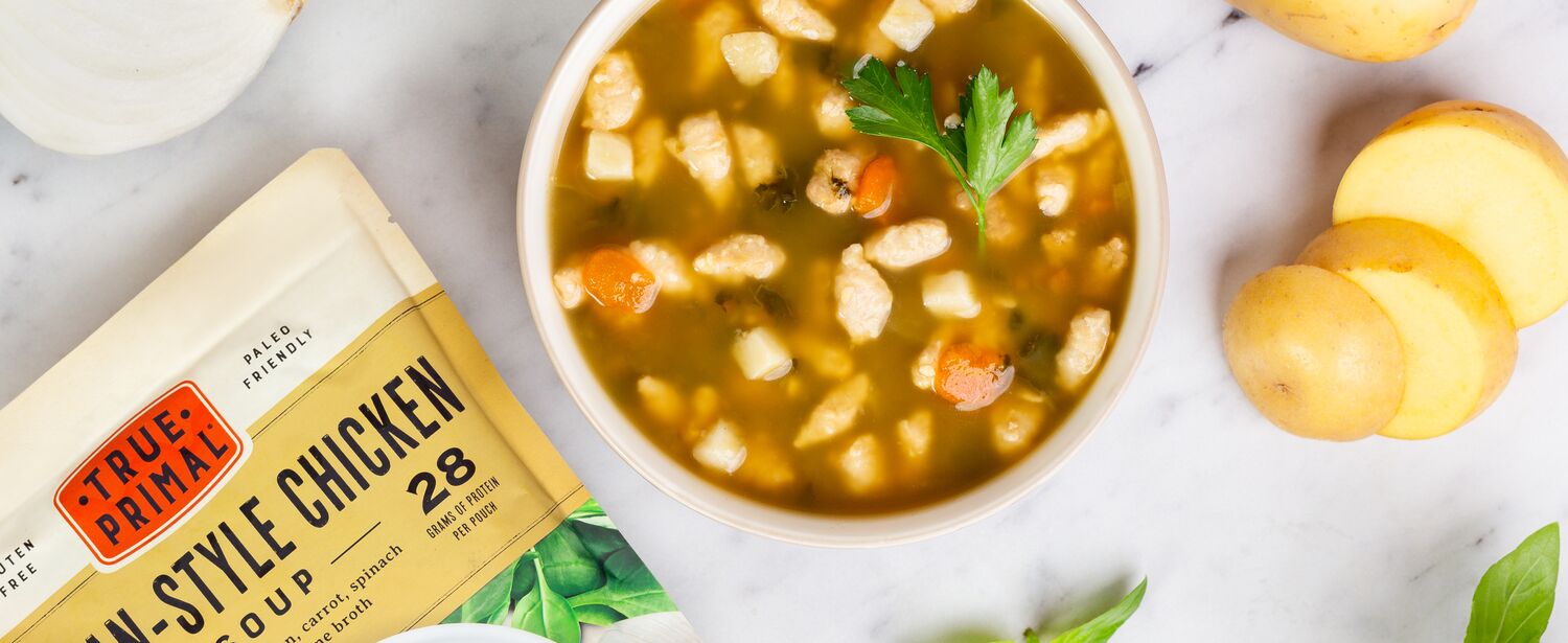 True Primal Tuscan-Style Chicken Soup in bowl next to pouch and vegetables