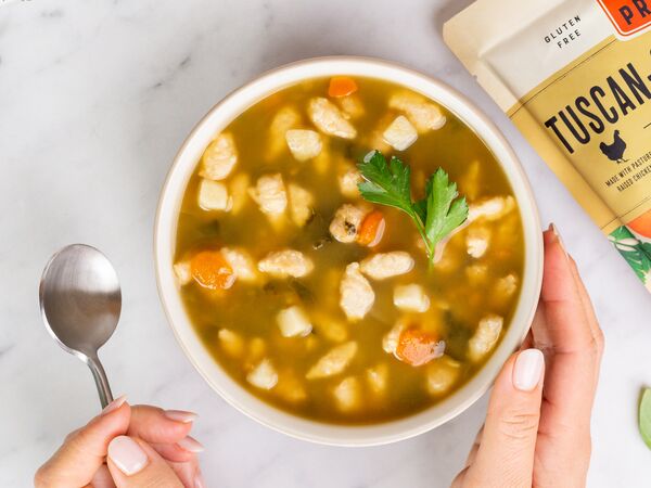 hand holding bowl of True Primal Tuscan-Style Chicken Soup with spoon near pouch