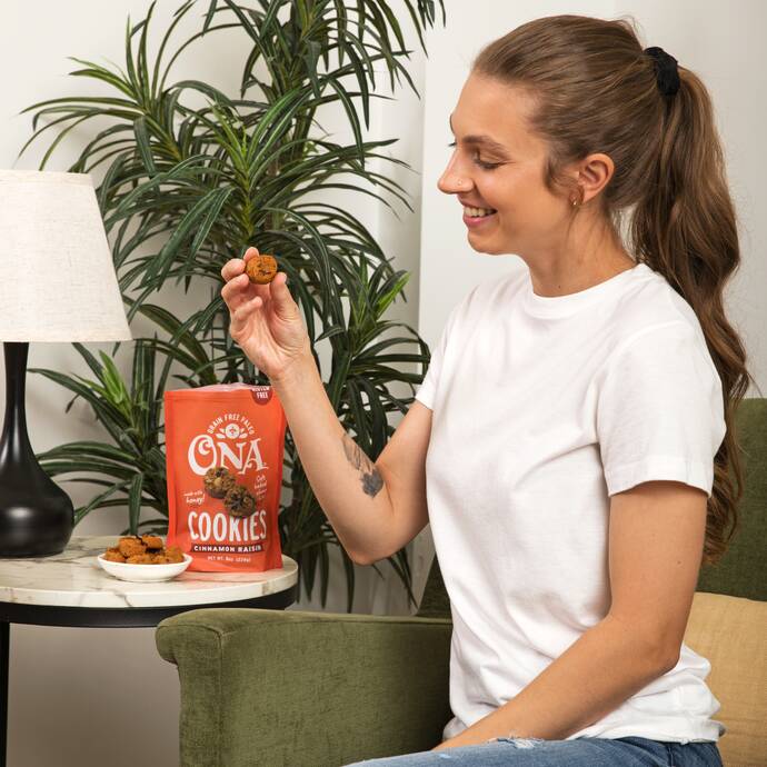 smiling woman on a couch eating Ona Ona Cinnamon Raisin Cookies